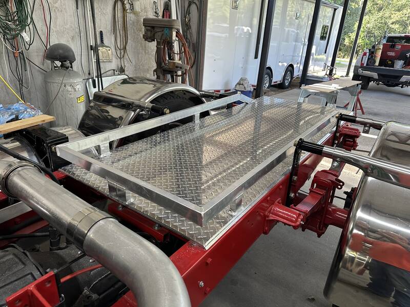 Aluminum diamond-plate deck with a tube frame mounted on a red truck chassis between chrome exhaust stacks, inside a shop