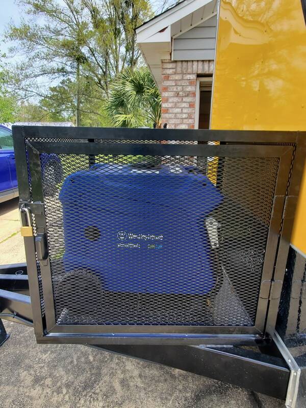 Black steel cage with an expanded-metal mesh door and a padlock, enclosing a blue portable generator on a trailer tongue