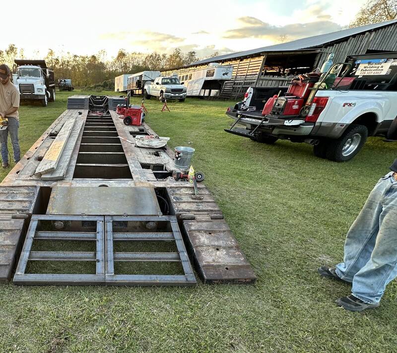 Lowboy trailer deck on grass with new bare-steel frame sections welded into its rear, tools on the deck and a pickup with a red welder parked alongside at golden hour