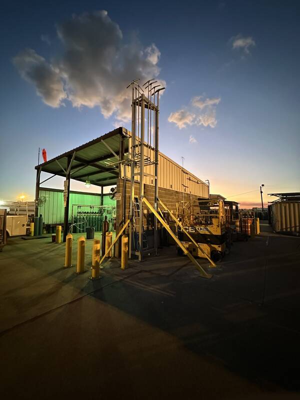 Tall galvanized steel frame tower with yellow diagonal braces beside a block building in an industrial yard, under a deep blue dusk sky