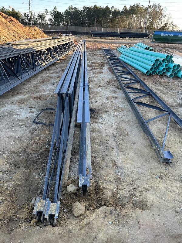 Long dark-gray steel trusses and beams laid out on bare dirt at a construction site, with green plastic pipe stacked to one side