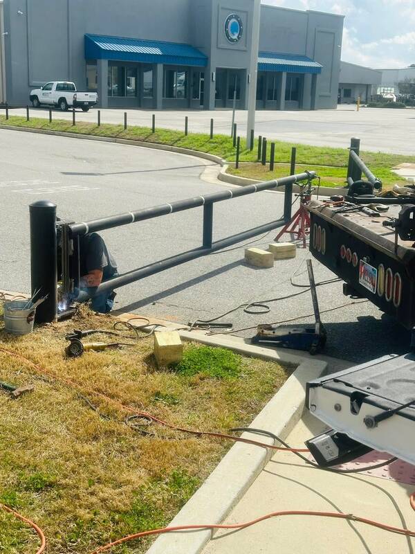 Welder crouched at a black steel post with the arc lit, welding a pipe barrier at the edge of a parking lot, with cables, a grinder and a bucket of rods on the grass and a work truck's flatbed at right